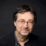 The Lions of Al-Rassan author Guy Gavriel Kay in dark attire, glasses, neutral background.