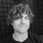 Legends & Lattes Trilogy author Travis Baldree with curly hair, black background.